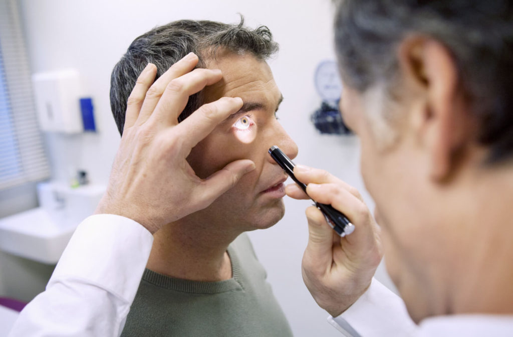 A male optometrist performing an eye exam for a male patient by shining a light toward the patient's eye to examine his eye health.