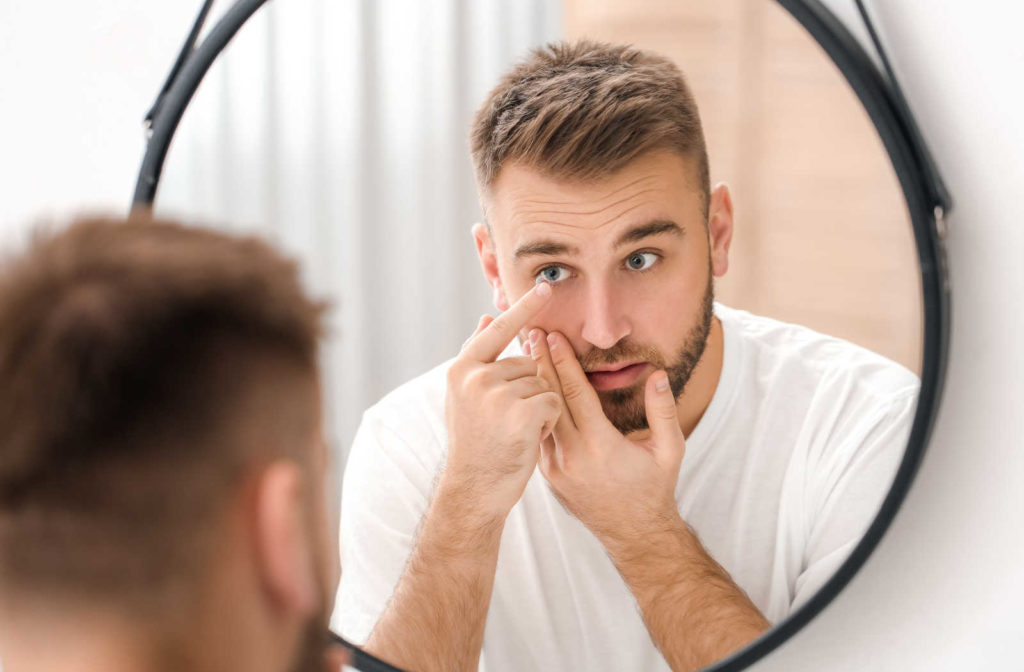 A man putting on a contact lens on his right eye using his right hand, while looking in the mirror.