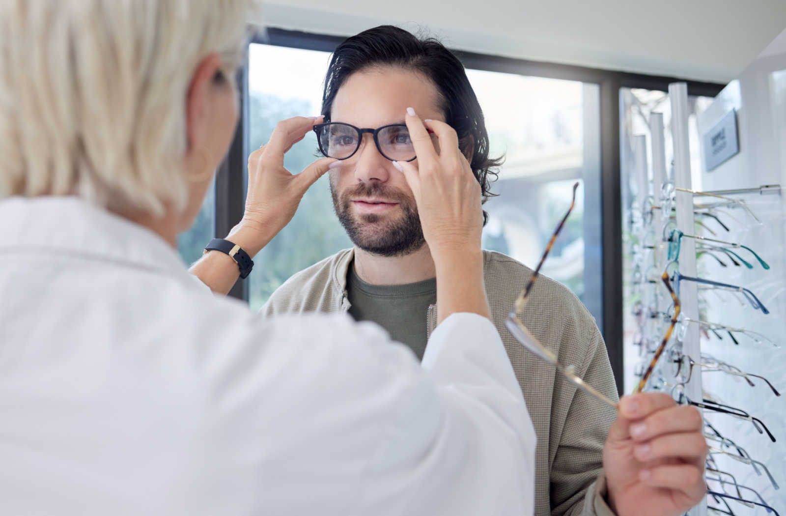 A man trying on glasses at an optic store while being assisted by an optometrist.