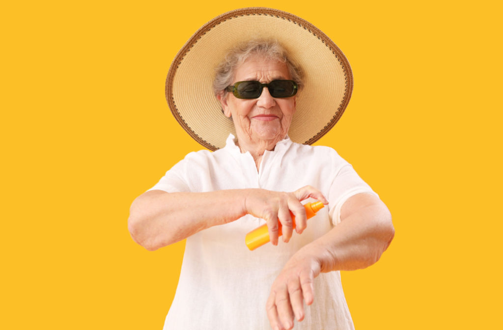An older woman wearing a wide-brimmed hat, dark sunglasses, and applying sunscreen to protect herself from UV radiation.